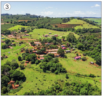 Imagem: Fotografia 3. Vista aérea de casas espalhadas entre árvores. Ao fundo, morros e mais árvores. Fim da imagem.