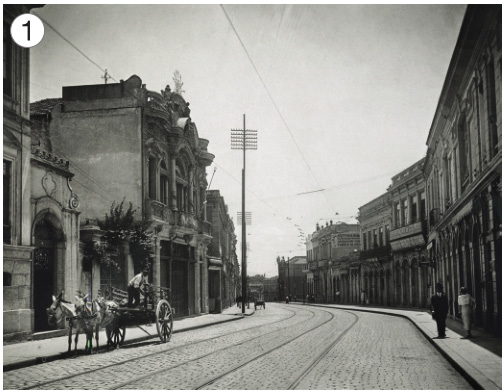 Imagem: Fotografia 1 em preto e branco. No centro há uma charrete em uma rua com um trilho no meio. Nas laterais há construções altas e postes. Fim da imagem.