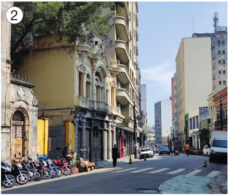 Imagem: Fotografia 2. No centro há várias motos estacionadas e carros em uma rua asfaltada com uma faixa de pedestres. Nas laterais há prédios e postes. Fim da imagem.