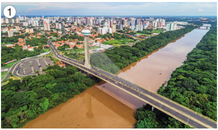 Imagem: Fotografia 1. Uma ponte sobre um rio com água marrom. Em volta há muitas árvores e ao fundo, uma cidade.  Fim da imagem.