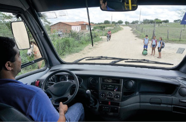 Imagem: Fotografia. Um homem está sentado e com as mãos no volante de um automóvel. Na frente dele, três crianças estão paradas. Ao fundo, uma casa e plantas.  Fim da imagem.