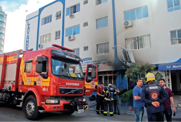 Imagem: Fotografia. Bombeiros estão ao lado de um caminhão vermelho. Ao fundo, um prédio com manchas pretas. Fim da imagem.