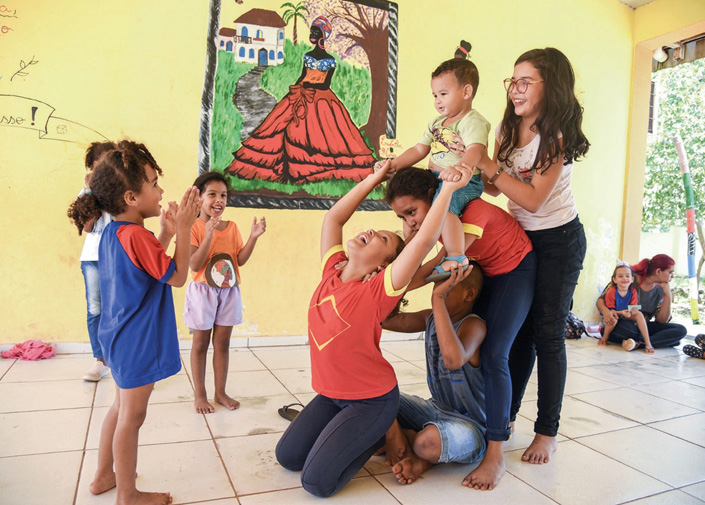 Imagem: Fotografia. À direita, uma menina está sorrindo e segurando um bebê, que está sentado nas costas de uma menina com o corpo inclinado para frente. Abaixo dela, um menino está sentado e segurando os pés do bebê. Na frente dele, uma menina está ajoelhada com o corpo inclinado para trás e segurando as mãos do bebê. Em volta, três crianças estão sorrindo e aplaudindo. Ao fundo, um quadro pendurado na parede e pessoas sentadas.  Fim da imagem.