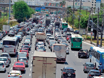 Imagem: Fotografia. Uma fileira de carros, caminhões e ônibus parados em uma avenida. Ao fundo, prédios e árvores. Fim da imagem.