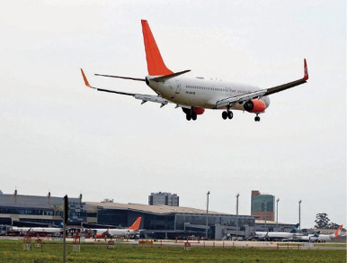 Imagem: Fotografia. Um avião pousando sobre uma pista. Ao fundo, o aeroporto.  Fim da imagem.