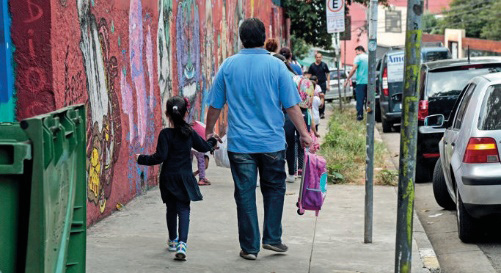 Imagem: Fotografia. Um homem está de costas, segurando a mão de uma menina com a mão esquerda e com a mão direita, ele segura uma mochila rosa. Eles estão andando na calçada e ao lado há uma fileira de carros.  Fim da imagem.