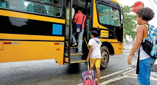 Imagem: Fotografia. Crianças estão enfileiradas e entrando em um ônibus escolar.  Fim da imagem.