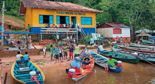 Imagem: Fotografia. Barcos coloridos atracados na frente de uma escola grande e amarela. Entre eles há várias pessoas. Fim da imagem.