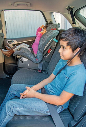 Imagem: Fotografia. Um menino está sentado no banco traseiro de um carro e com cinto de segurança em volta do corpo. Ao seu lado, uma menina está sentada em um cadeirão com cinto de segurança.  Fim da imagem.