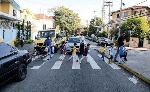 Imagem: Fotografia. Várias crianças estão enfileiradas e atravessando uma rua sobre a faixa de pedestres. Entre elas há dois adultos e atrás da faixa há carros parados. Ao fundo, casas e árvores. Fim da imagem.