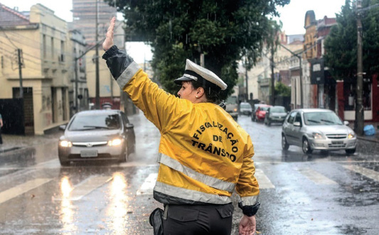 Imagem: Fotografia. Uma pessoa com quepe branco, casaco amarelo com a informação: FISCALIZAÇÃO DE TRÂNSITO e calça preta está de costas com a mão esquerda levantada. Na frente dela há carros parados atrás de uma faixa de pedestres e em volta há chuva caindo. Ao fundo, árvores e prédios. Fim da imagem.