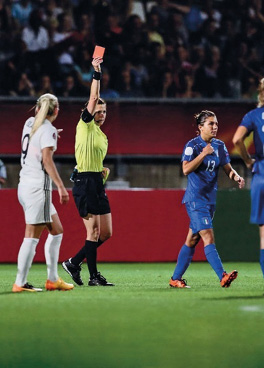 Imagem: Fotografia. No centro, uma mulher com camiseta verde, bermuda e chuteiras pretas está segurando um cartão vermelho com a mão direita levantada. Na frente dela, uma jogadora com uniforme azul está andando e ao lado, uma jogadora com uniforme branco observa. Ao fundo, pessoas sentadas em uma arquibancada. Fim da imagem.