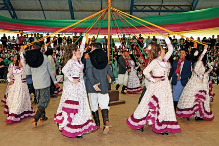 Imagem: Fotografia. Mulheres com vestido branco e rosa estão com as mãos direitas na cintura e segurando fitas amarelas com a mão esquerda levantada. As fitas estão presas em uma haste no centro. Entre as mulheres há homens com chapéu preto pendurado no pescoço, camisa cinza, calça branca e botas pretas. Eles estão com as mãos esquerdas atrás das costas. Ao fundo, uma multidão observa sentada em uma arquibancada. Fim da imagem.