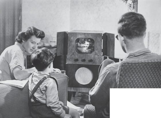 Fotografia em preto e branco. Um casal e um menino estão sentados e na frente deles há uma televisão com a tela pequena, dentro de um tubo de madeira grande e com portas nas laterais. 