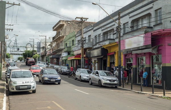 Imagem: Fotografia. No centro há uma rua com carros. Ao lado, pessoas andando na calçada, na frente de lojas. Ao fundo, prédios e nuvens no céu cinza.  Fim da imagem.