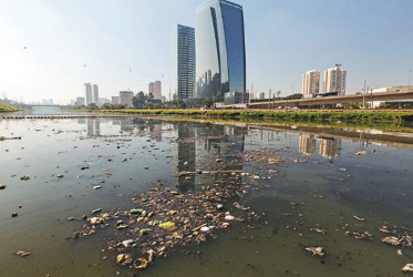 Imagem: Fotografia. Um rio com água escura e lixo boiando. Ao fundo, prédios e construções.  Fim da imagem.