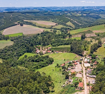 Imagem: Fotografia. Vista aérea de uma região montanhosa. À direita há várias casas e à esquerda, morros e árvores. Fim da imagem.