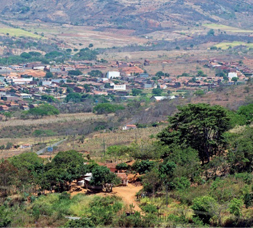 Imagem: Fotografia. Em primeiro plano há várias árvores. No centro, casas espalhadas e ao fundo, morros e árvores.  Fim da imagem.