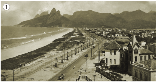 Imagem: Fotografia 1 em preto e branco. À esquerda, a orla da praia. No centro, uma rua com dois carros e uma charrete. À direita, casas e árvores. Ao fundo, morros. Fim da imagem.