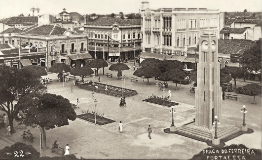 Imagem: Fotografia em preto e branco. Uma praça com bancos e árvores no chão de terra. No centro há uma torre com um relógio na ponta. Em volta há várias construções e pessoas andando. Fim da imagem.