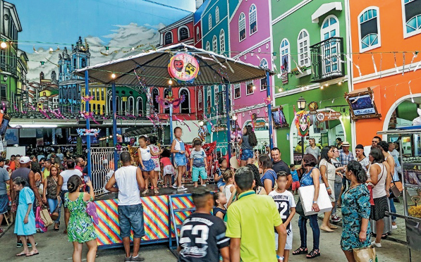 Imagem: Fotografia. No centro há crianças em um coreto. Em volta há várias pessoas e nas laterais, casas coloridas.  Fim da imagem.