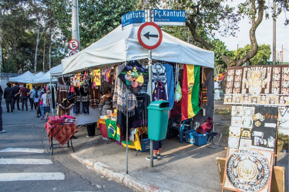 Imagem: Fotografia. No centro, destaque para uma tenda com produtos coloridos. Ao lado, uma rua com faixa de pedestre e ao fundo, mais tendas e árvores. Fim da imagem.