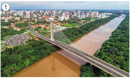 Imagem: Fotografia 1. Uma ponte sobre um rio com água marrom. Em volta há muitas árvores e ao fundo, uma cidade.  Fim da imagem.