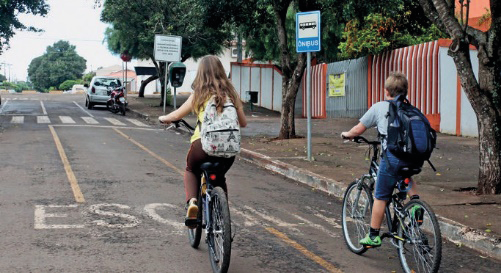 Imagem: Fotografia. Duas crianças com mochilas nas costas estão andando de bicicleta.  Fim da imagem.