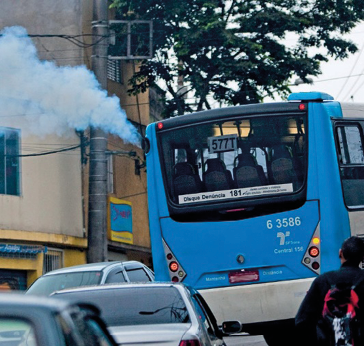 Imagem: Fotografia. Um ônibus azul está andando e soltando fumaça. Em volta há vários carros e ao fundo, construções. Fim da imagem.