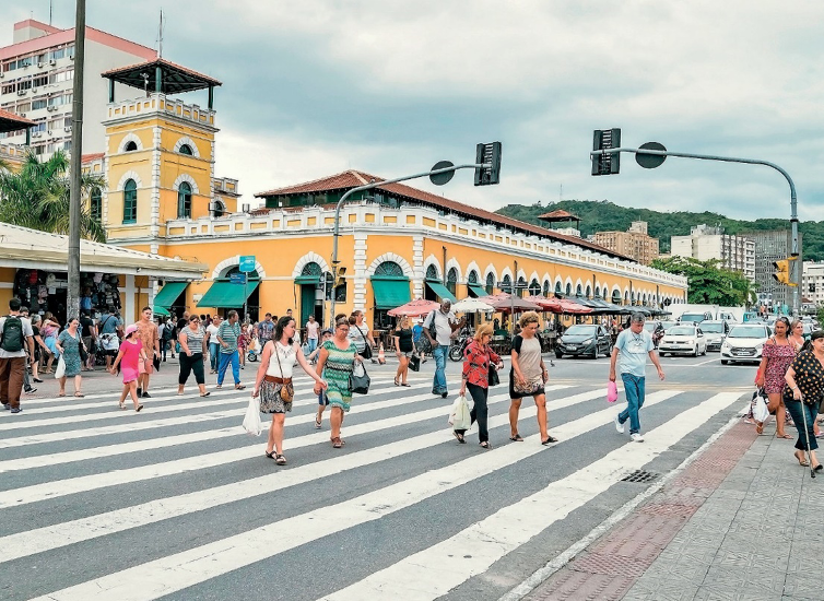 Imagem: Fotografia. Várias pessoas estão atravessando uma rua sobre uma faixa de pedestres. Atrás delas há carros parados e ao fundo, uma construção extensa com paredes amarelas. Fim da imagem.