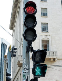 Imagem: Fotografia. Na parte superior, um semáforo com a luz vermelha acesa. Na parte inferior, um semáforo de pedestres com a luz verde acesa.  Fim da imagem.