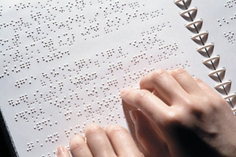 Imagem: Fotografia. Mãos de uma pessoa sobre um livro com textos em Braille. Fim da imagem.