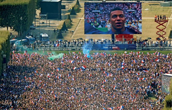 Imagem: Fotografia. Vista aérea de uma multidão aglomerada em um campo. Na frente há um telão pendurado com a imagem de um jogador com uniforme azul. Atrás dele há várias pessoas sentadas em uma arquibancada. Fim da imagem.