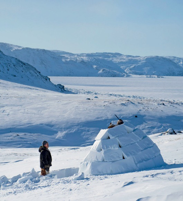 Imagem: Fotografia. Uma pessoa com gorro e blusa está em pé com as pernas na neve. Na frente dela há um iglu feito de blocos de neve. Ao fundo, morros cobertos de neve. Fim da imagem.