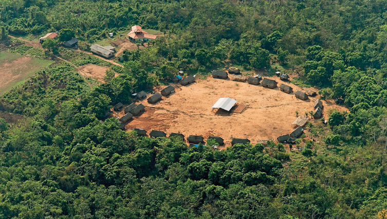 Imagem: Fotografia. Vista aérea de uma floresta. No centro há uma área desmatada com ocas formando um círculo em volta de um galpão grande com telhado.  Fim da imagem.