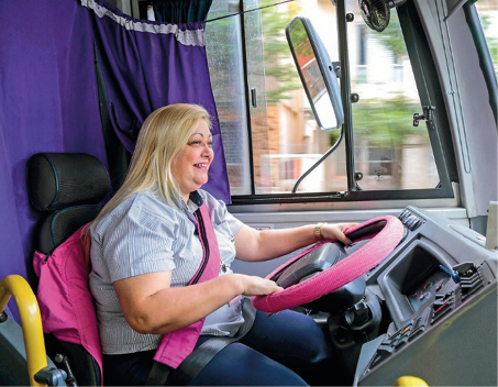 Imagem: Fotografia. Uma mulher loira com camisa cinza e calça azul está sorrindo e com as mãos sobre um volante rosa. Atrás dela há uma cortina roxa. Fim da imagem.