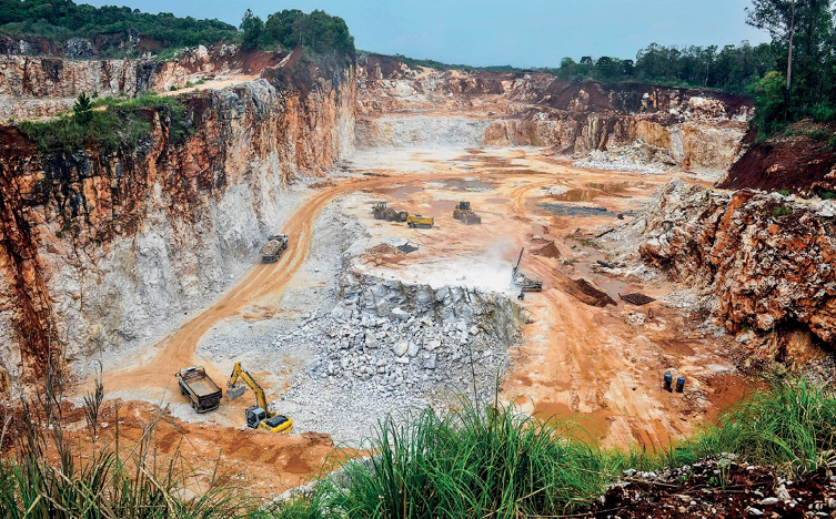 Imagem: Fotografia. Caminhões em um terreno com pedras. Em volta há paredes de terra. Fim da imagem.