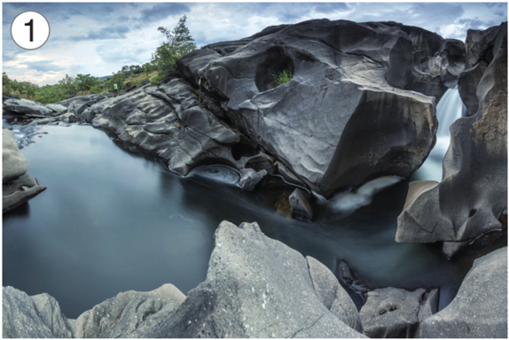 Imagem: Fotografia 1. À direita, uma cachoeira com rochas em volta. À esquerda, um rio e ao fundo, árvores.  Fim da imagem.
