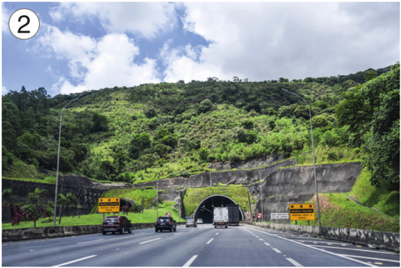 Imagem: Fotografia 2. Carros sobre uma estrada e no centro há um túnel em um morro com árvores. Fim da imagem.