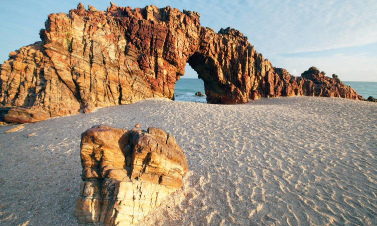 Imagem: Fotografia. No centro há uma formação rochosa em tons de marrom com um buraco no meio e sobre areia clara. Ao fundo, o mar. Fim da imagem.