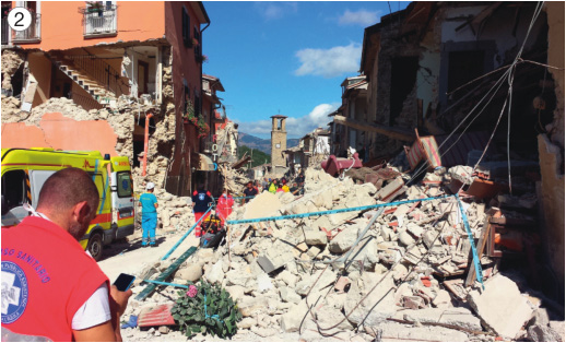 Imagem: Fotografia 2. Pessoas em volta de entulhos e ao lado há construções com paredes quebradas.  Fim da imagem.