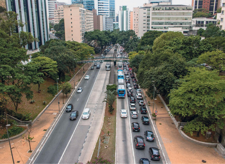 Imagem: Fotografia. Vista aérea de uma avenida. À esquerda há menos veículos e à direita, uma fileira de automóveis parados. Nas laterais há várias árvores e ao fundo, prédios. Fim da imagem.