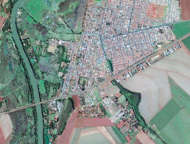 Imagem: Fotografia. Vista de cima de uma cidade com muitas construções. Em volta há campos verdes e árvores. À esquerda, um rio extenso. Fim da imagem.