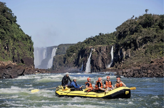 Imagem: Fotografia. Seis pessoas com capacete e colete estão sentadas em um bote inflável amarelo em um rio e segurando remos. Ao fundo há várias cachoeiras e árvores. Fim da imagem.
