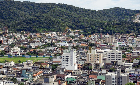 Imagem: Fotografia. Vista aérea de várias casas e prédios. Ao fundo, um morro coberto de árvores. Fim da imagem.