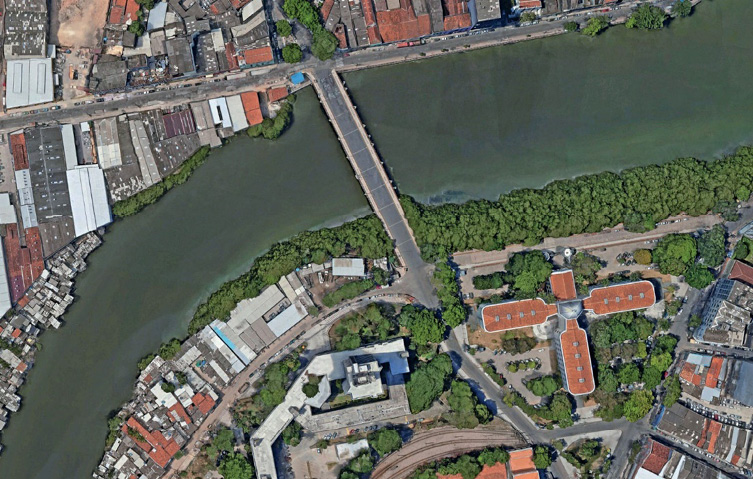 Imagem: Fotografia. Vista de cima de um rio sinuoso. No centro há uma ponte e nas margens há árvores, construções e ruas. Fim da imagem.