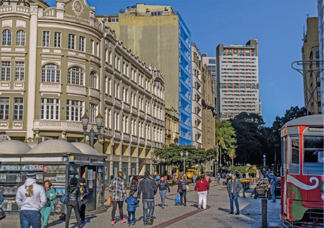 Imagem: Fotografia. No centro há pessoas andando na calçada. À esquerda, prédios e construções e à direita, um bonde na rua. Fim da imagem.