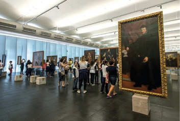 Imagem: Fotografia. Pessoas observando pinturas penduradas em um salão grande.  Fim da imagem.