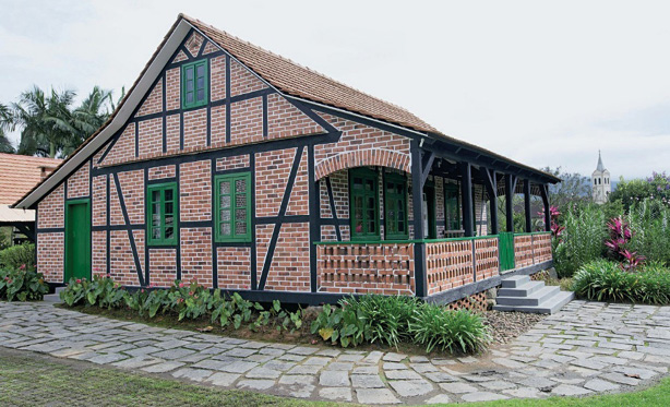 Imagem: Fotografia. Casa com paredes de tijolos e detalhes em verde. Em volta há plantas e flores.  Fim da imagem.