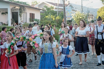 Imagem: Fotografia. Meninas com vestido azul e vermelho estão segurando arcos de flores coloridas. Ao fundo, várias pessoas, casas e árvores. Fim da imagem.